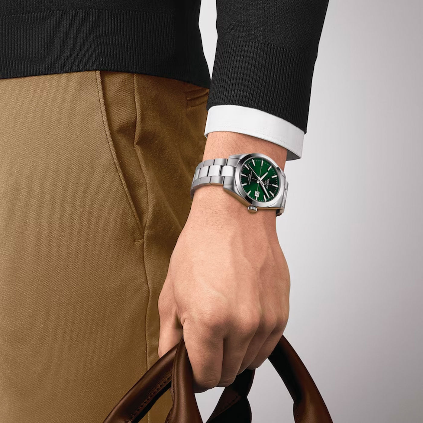 A person wearing a black sweater with a white cuff holds a brown leather bag. On their left wrist, they have the Tissot Gentleman Powermatic 80 Automatic Silicium 40mm Watch, which showcases the elegance of Swiss Automatic Movement with its silver construction and green dial. The background is plain and neutral.