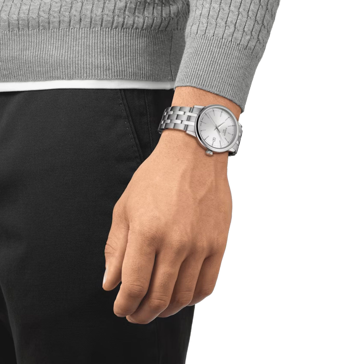 A person dressed in a gray sweater and black pants is sporting the Tissot Classic Dream Quartz 42mm Watch by Tissot on their left wrist. The watch features a scratch-resistant sapphire crystal, a metallic band, and a round, light-colored face with minimal design. The background is plain white.