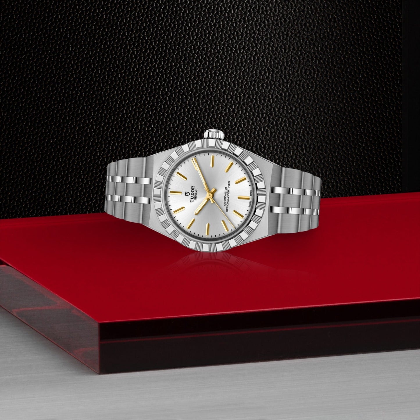 A TUDOR Royal wristwatch by TUDOR, featuring a silver case, white dial, and gold hour markers, rests on a red surface against a textured black background—a stylish embodiment of automatic sport-chic timepieces.