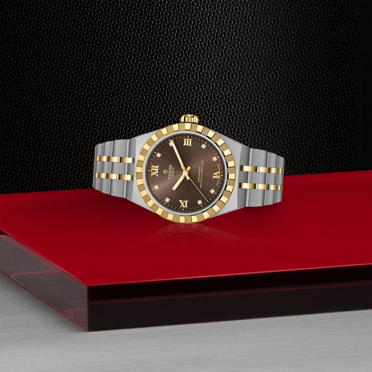 The TUDOR Royal by TUDOR features a two-tone gold and silver metal band and a black face with gold Roman numerals, shown on a sleek red and gray surface against a textured black background.