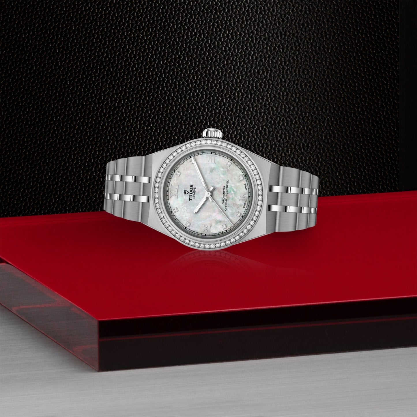 A TUDOR Royal wristwatch by TUDOR, featuring a silver case, mother-of-pearl dial, and diamond bezel with integrated bracelet, is showcased on a glossy red and black platform against a textured black background.