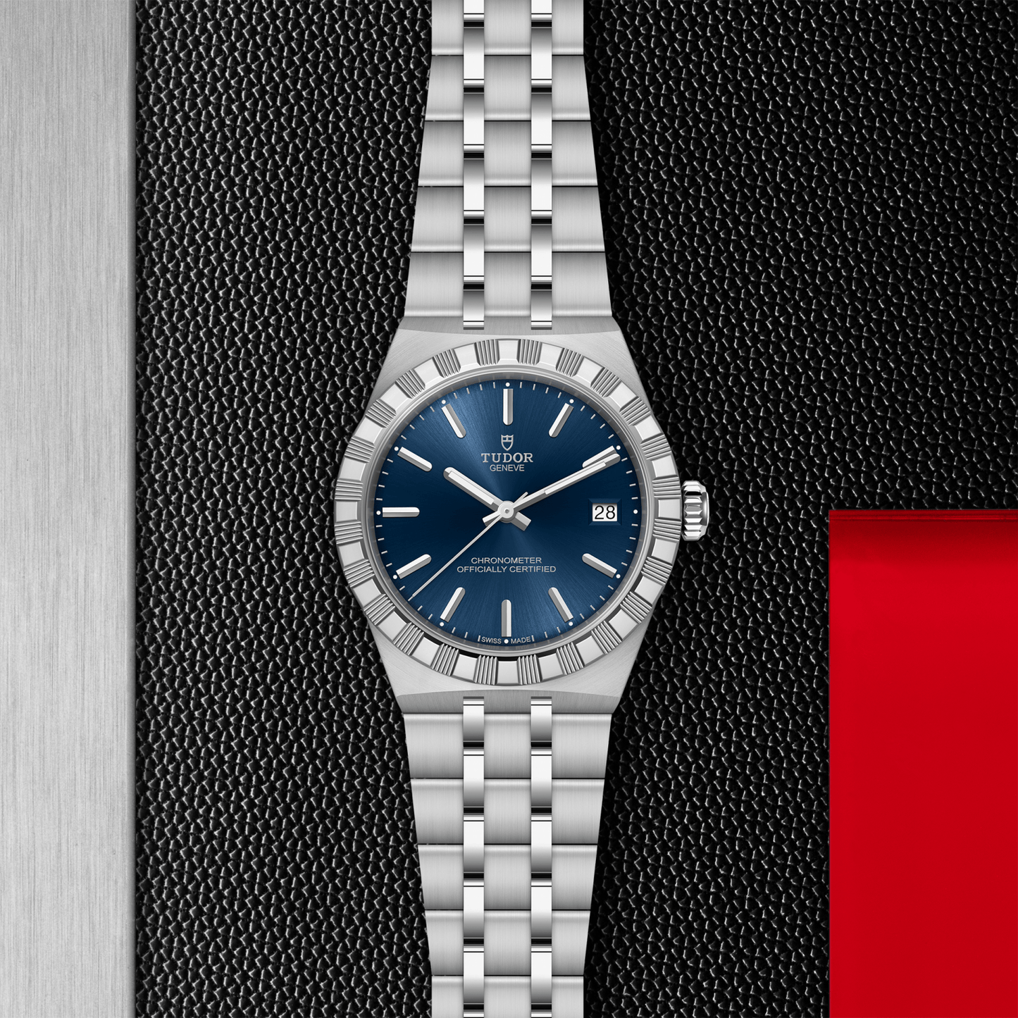 The TUDOR Royal by TUDOR features a stainless steel case and bracelet, a blue dial with silver hour markers, and a date window at 3 o’clock, set against a textured black background with silver and red accents.