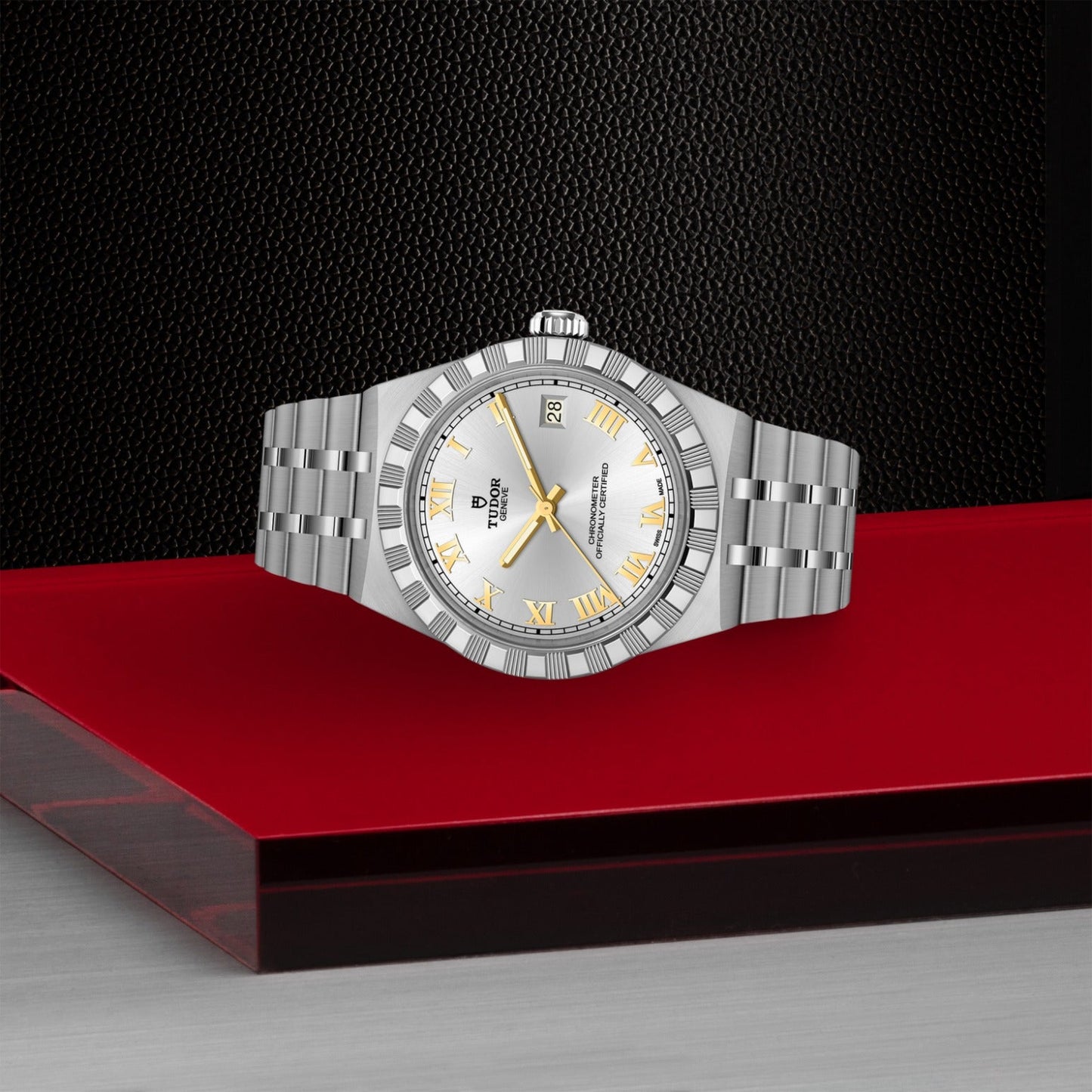 A TUDOR Royal watch by TUDOR, featuring a silver case, white dial, gold Roman numerals, and date display, is displayed on a red platform against a black textured backdrop—exemplifying automatic sport-chic elegance.