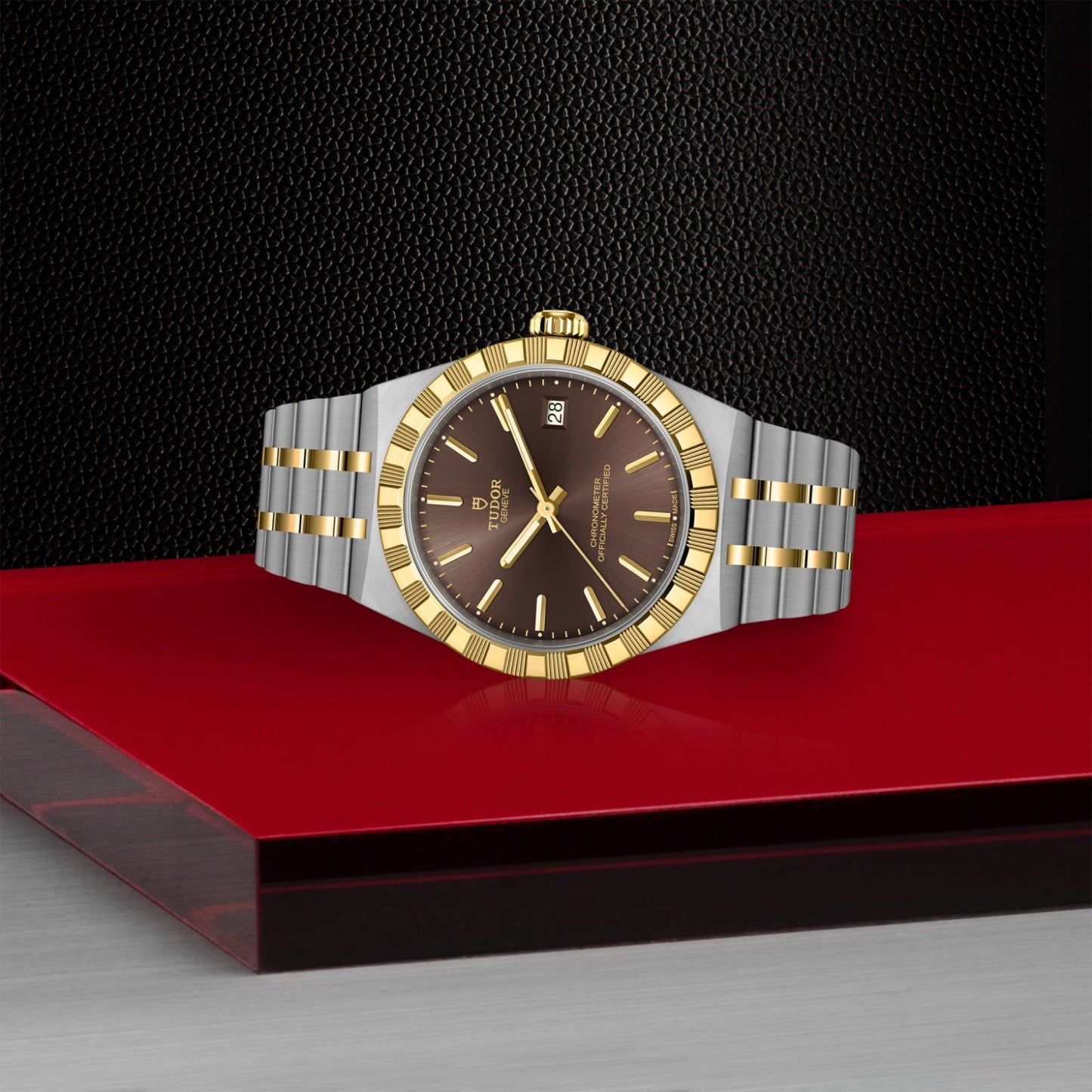The TUDOR Royal by TUDOR, featuring a silver and gold metal band, black dial, gold bezel, and gold markers, is showcased on a glossy red surface with a dark textured background—highlighting its automatic sport-chic style.