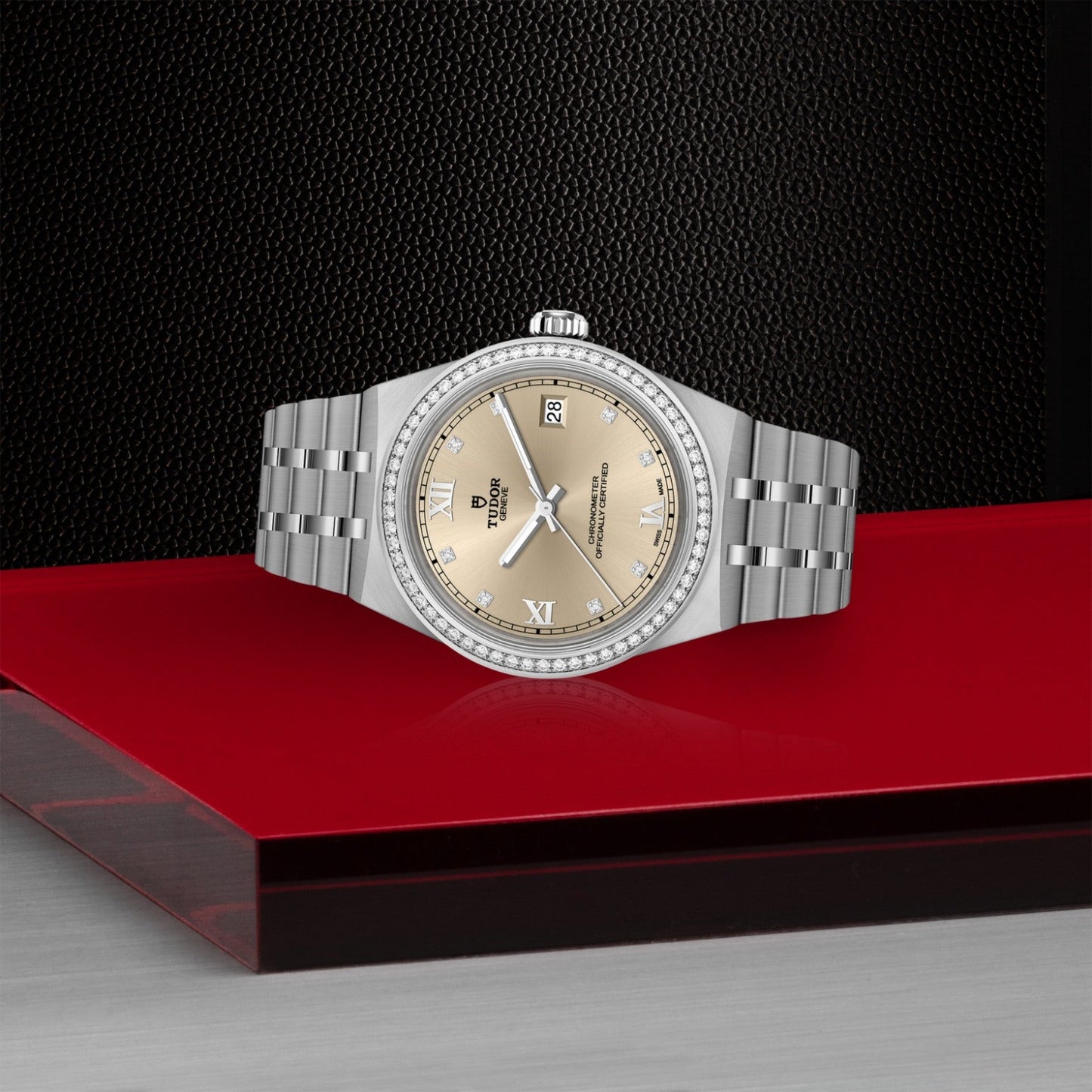 The TUDOR Royal by TUDOR, a silver luxury wristwatch with a champagne dial, Roman numerals, and a diamond-set bezel, is showcased on a red and black platform against a textured black background.