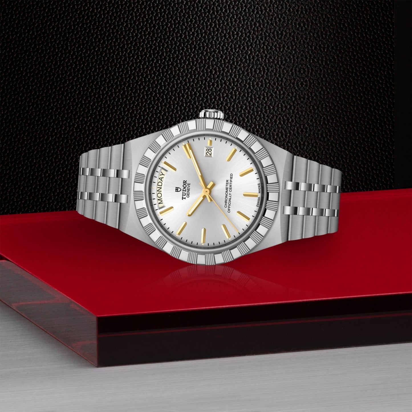 The TUDOR Royal by TUDOR, featuring a silver case, white dial with gold hour markers, and a metal link band, is shown on a red and gray surface against a black textured background. This automatic watch displays the day and date.