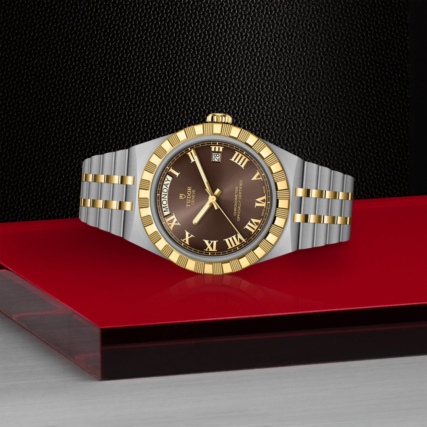 A TUDOR Royal luxury wristwatch by TUDOR, featuring a gold and silver band, brown dial with gold Roman numerals, and day/date display—an elegant automatic sport-chic watch shown on a red and silver surface against textured black.