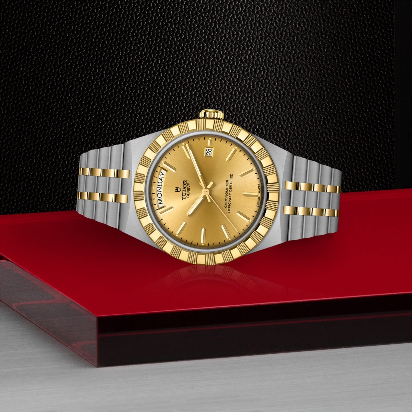 The TUDOR Royal, a luxury wristwatch by TUDOR with a gold dial and bezel, two-tone stainless steel bracelet, and day-date display, rests on a glossy red and silver surface against a textured black background.