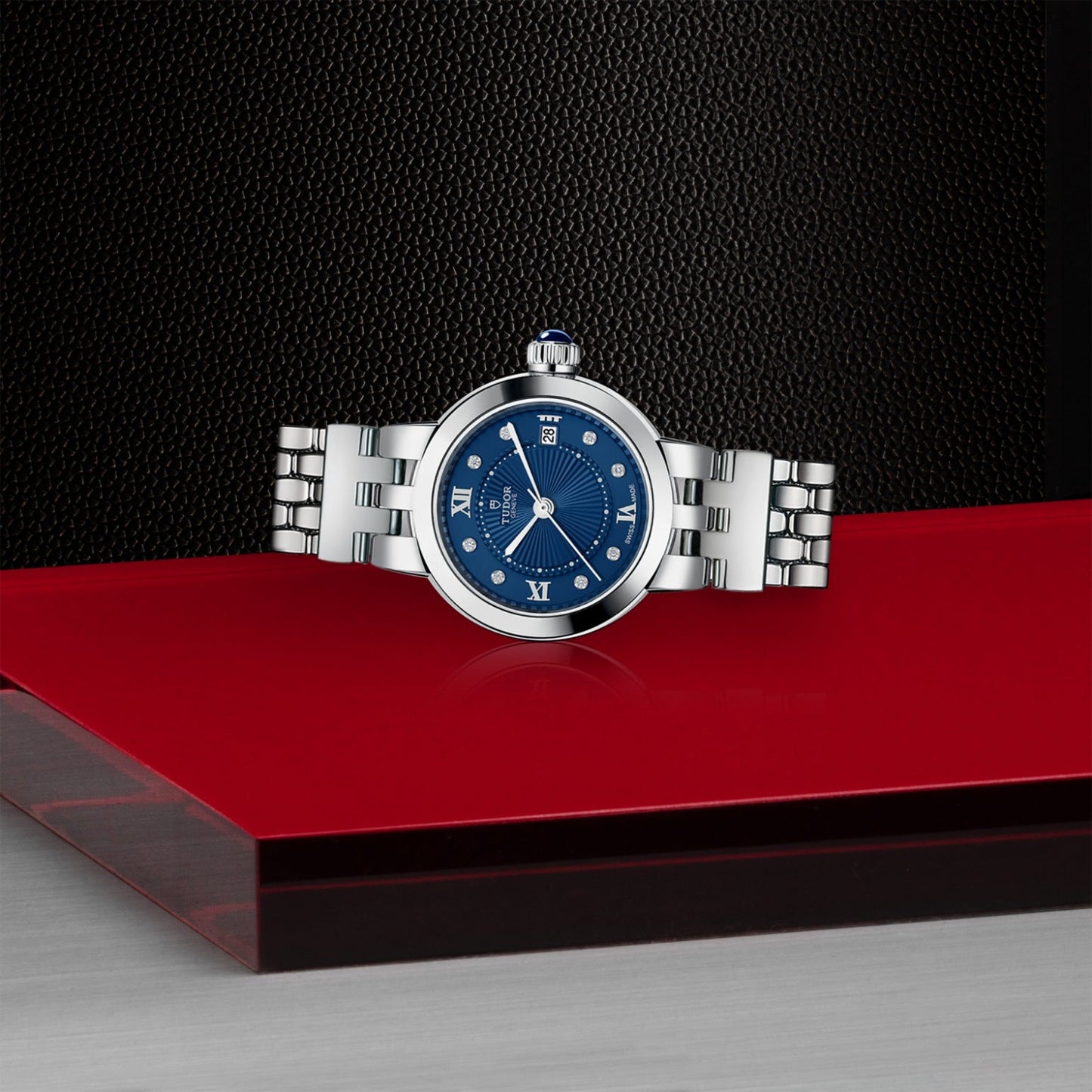 The TUDOR Clair de Rose, featuring a steel bracelet and opaline blue dial with Roman numerals, is showcased on a glossy red platform set against a textured black background.
