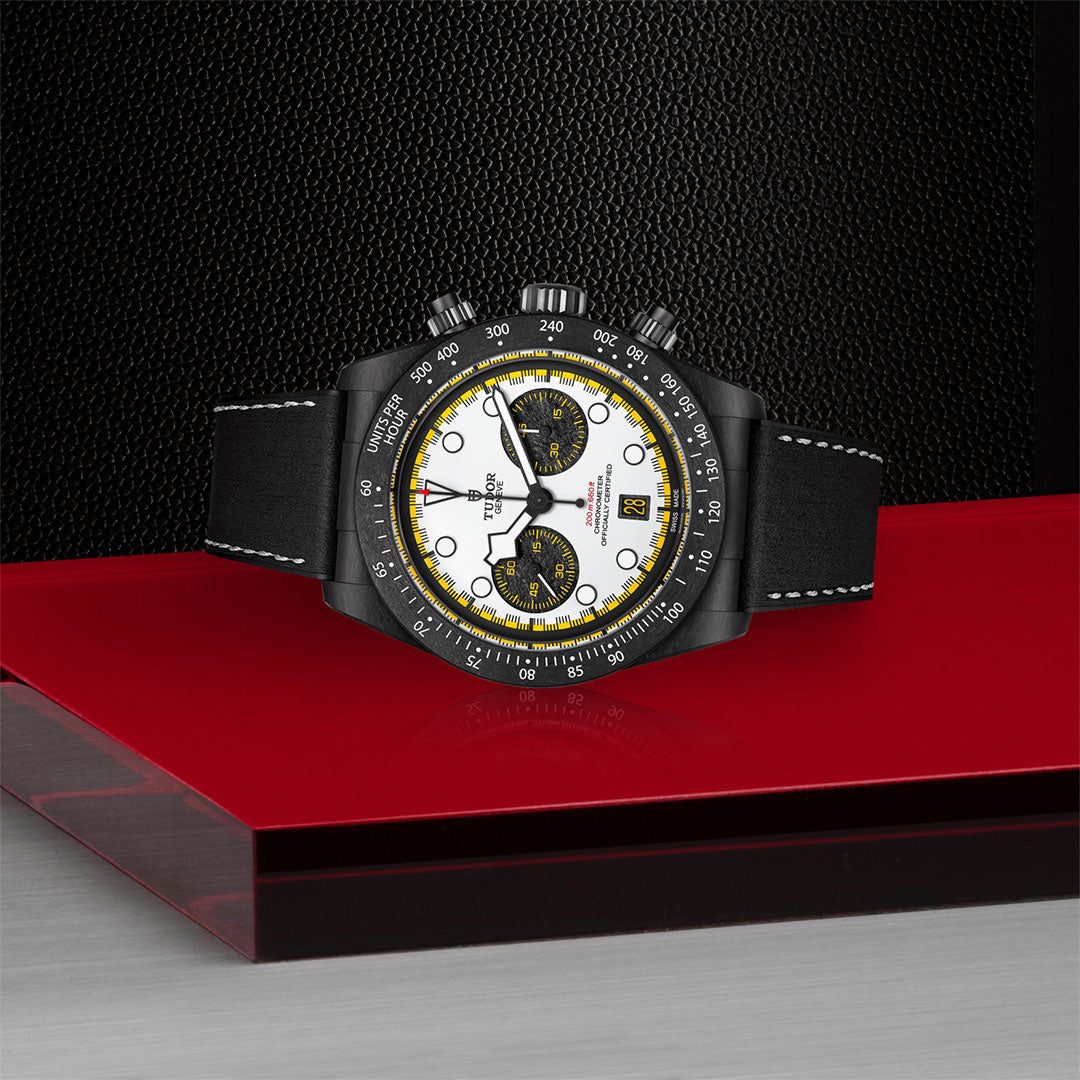 The TUDOR Black Bay Chrono, featuring a black leather strap, white dial, and yellow accents, is showcased on a glossy red and silver surface against a textured black background.
