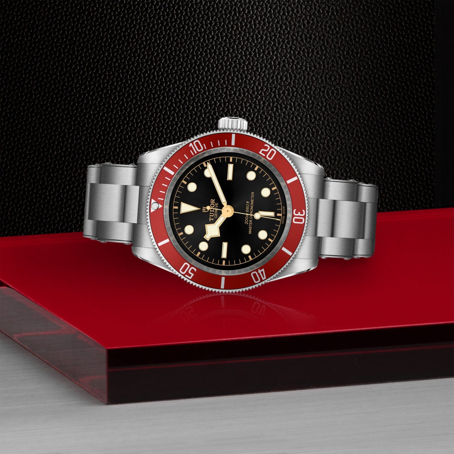 The TUDOR Black Bay by TUDOR features a 41mm steel case, stainless steel band, black dial with bold white markers, red bezel, and gold accents—shown on a red and metallic surface set against a textured black background.