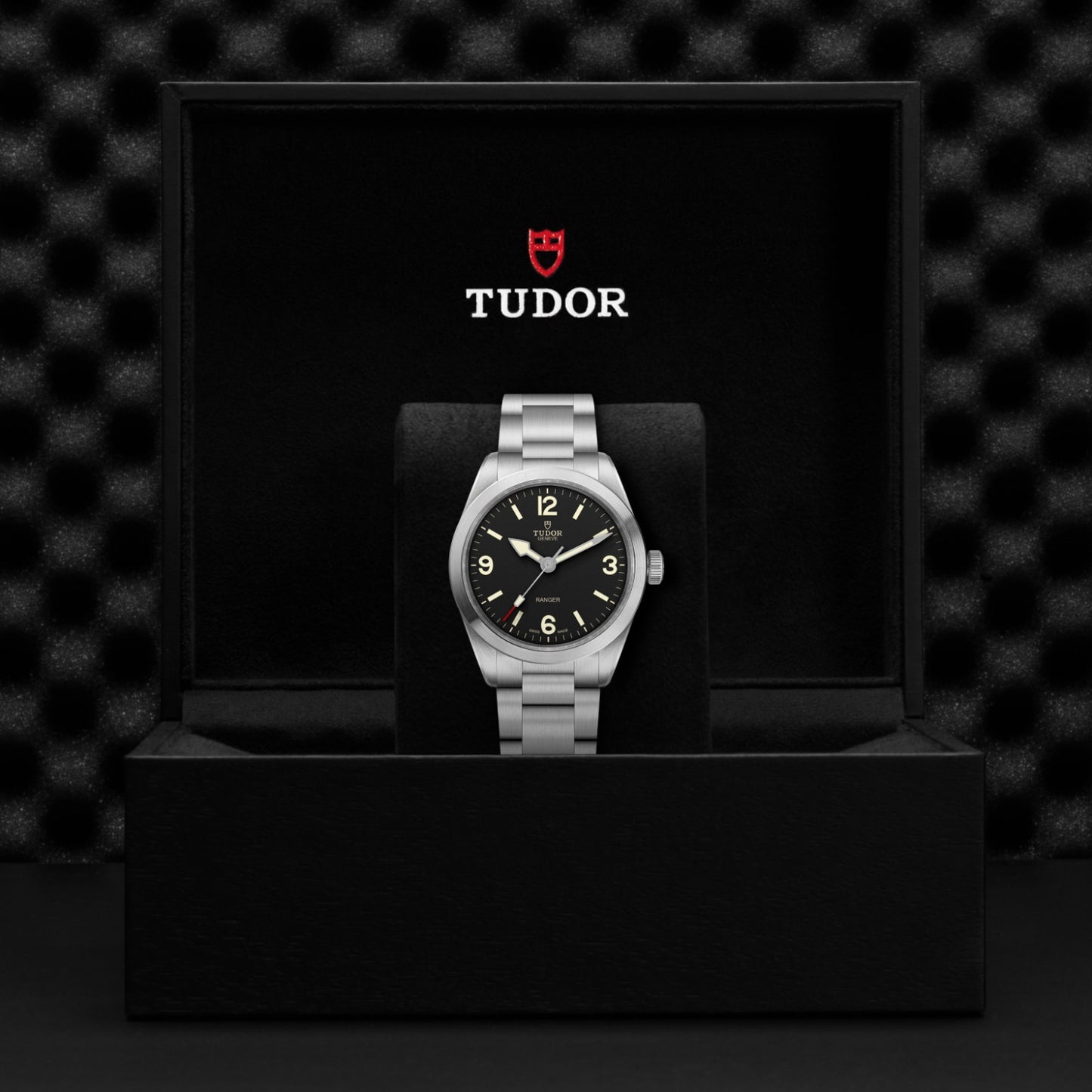 The TUDOR Ranger watch, featuring a 39mm stainless steel case and black dial, is shown in an open black box with the TUDOR logo in white and red on the lid, set against a textured black background.