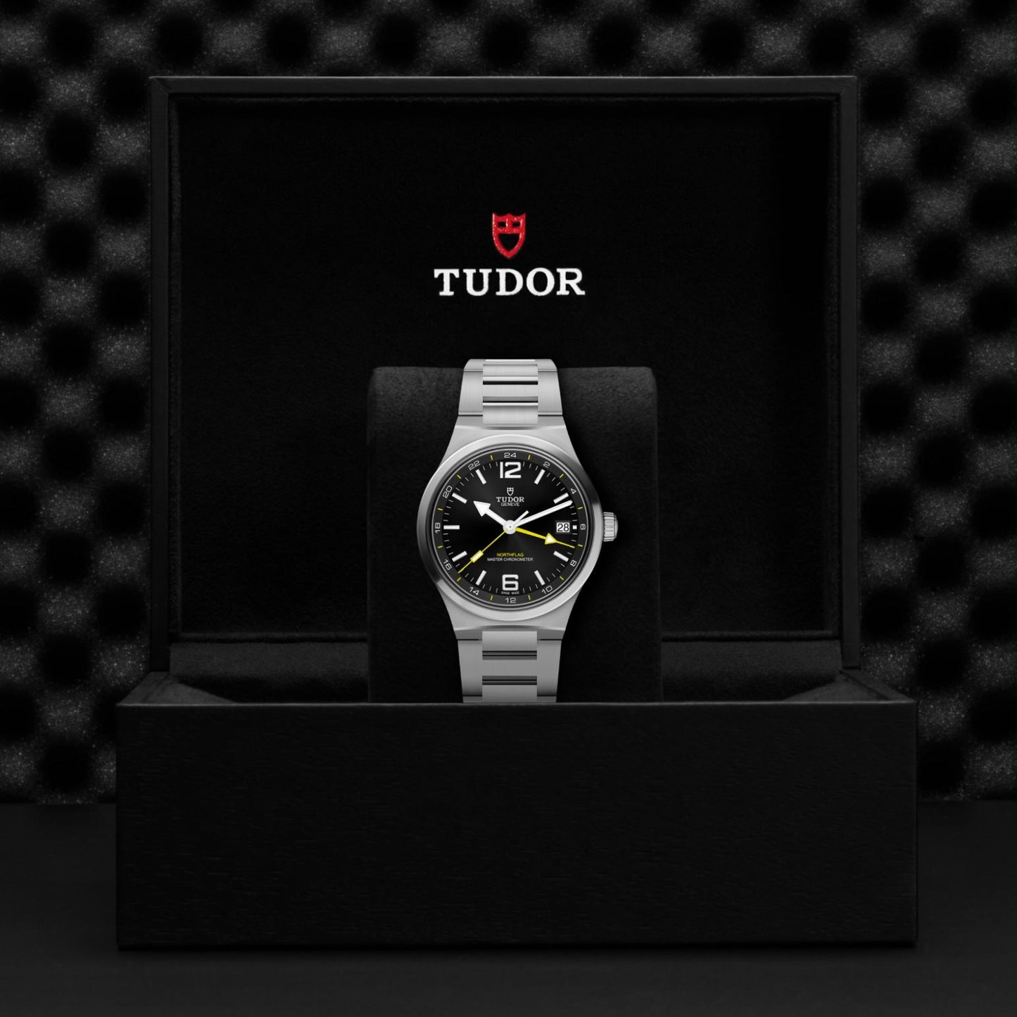 A TUDOR North Flag wristwatch with a silver band is displayed in an open black box featuring the TUDOR logo and name above it. With Master Chronometer certification, the watch showcases a black face with white and yellow details inspired by the iconic TUDOR North Flag design. The background is dark and textured.