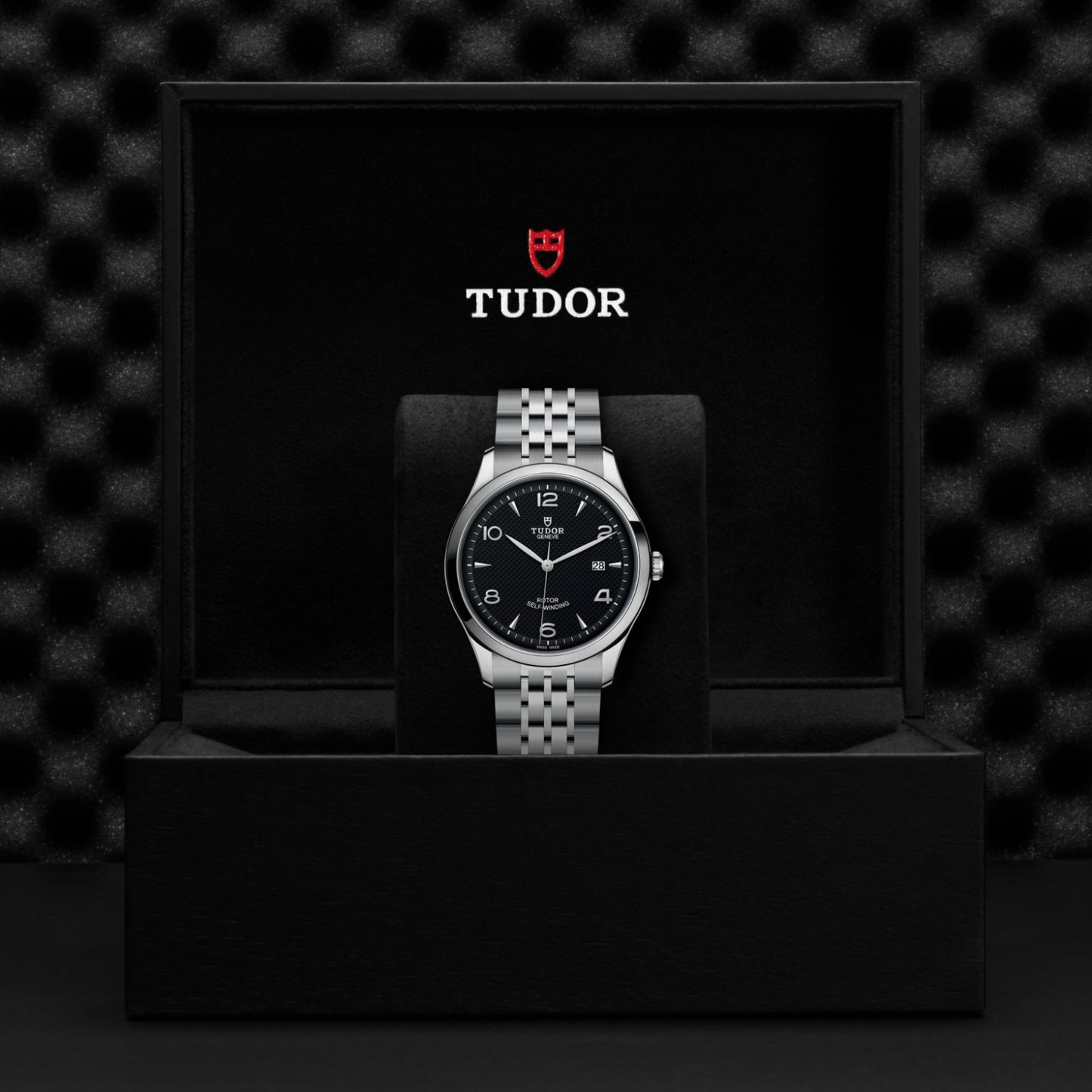 A silver TUDOR 1926 wristwatch with a black dial is showcased upright in an open black TUDOR box against a textured black background, highlighting the brand’s timeless elegance.
