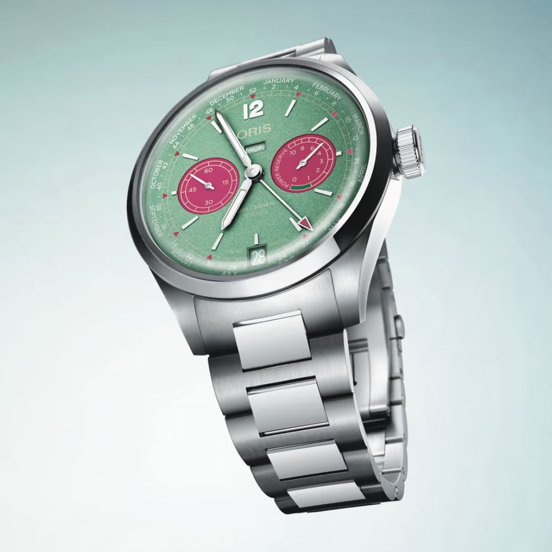 The ORIS Big Crown Calibre 113 Automatic Winding 43mm Watch features a silver case, green dial with two red subdials, stainless steel bracelet, and business calendar functions—all set against a light blue gradient background.