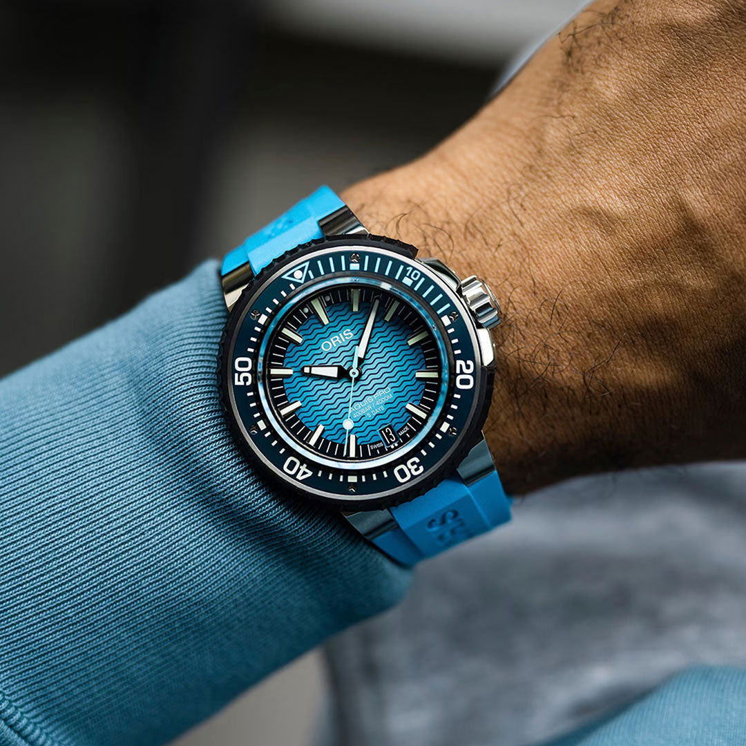 An individual wears the ORIS Aquis Pro 4000m Automatic Winding 49.5mm Watch, featuring a wave-patterned blue dial and matching blue strap, on their wrist against a blue sleeve. This remarkable timepiece from ORIS is engineered for incredible durability, offering an exceptional 4000-metre water resistance.