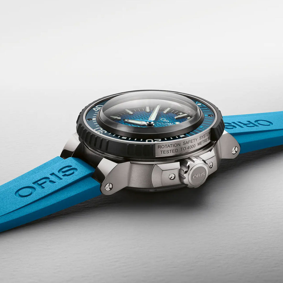 A close-up of the ORIS Aquis Pro 4000m Automatic Winding 49.5mm Watch showcases its blue strap, black bezel, and metallic silver case. The watch face displays striking blue accents, while the side features a label reading “Rotation Safety System Tested to 4000 meters,” emphasizing its remarkable 4000-metre water resistance as an advanced diver’s timepiece.