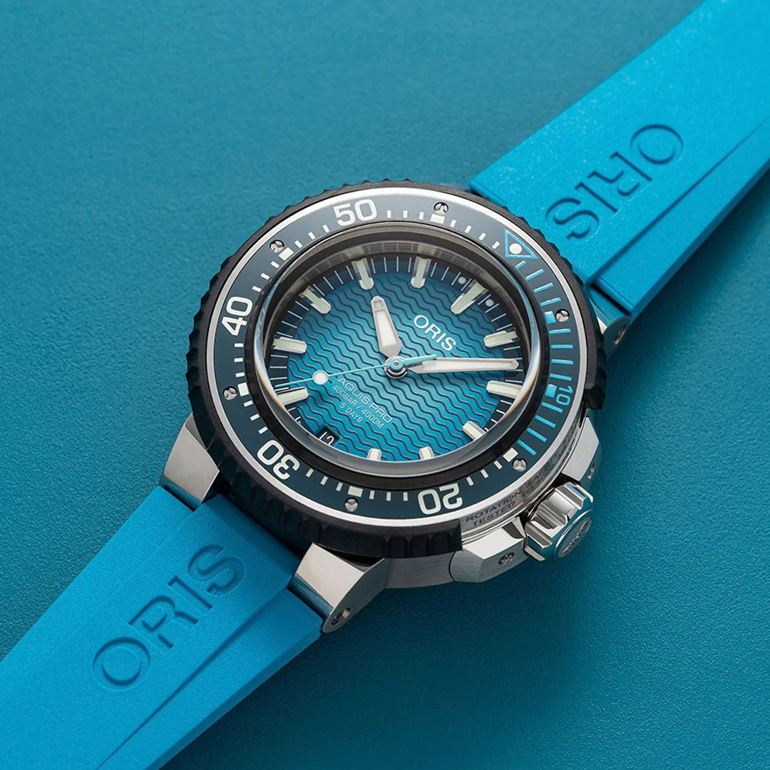 A close-up of the ORIS Aquis Pro 4000m Automatic Winding 49.5mm Watch features its striking blue rubber strap, wave-patterned dial, and bold white markers, set against a blue surface—highlighting the watch’s impressive 4000-metre water resistance.
