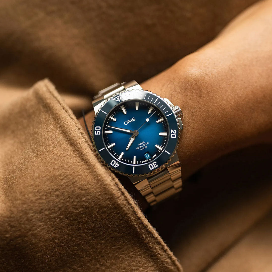 A wrist wearing the ORIS Aquis Date Calibre 400 Automatic Winding 43.5mm Watch with blue dial and bezel, partly hidden by a tan coat sleeve, highlights the precision engineering of ORIS.