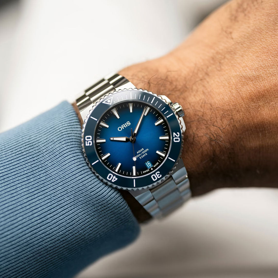 A person in a blue long-sleeve shirt displays the ORIS Aquis Date Calibre 400 Automatic Winding 43.5mm Watch, featuring a metallic strap, blue gradient dial with white markers, and unidirectional bezel—a true luxury toolwatch.