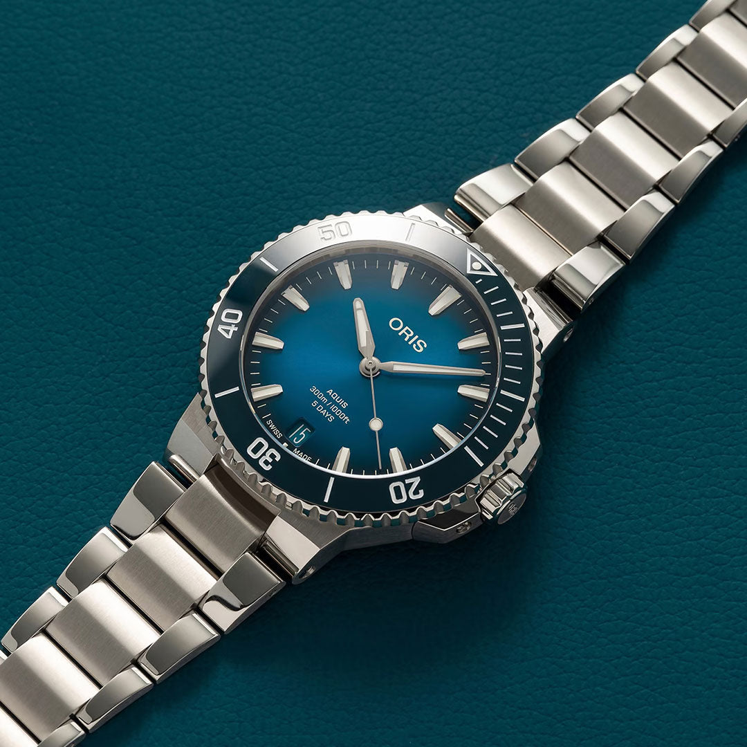 The ORIS Aquis Date Calibre 400 Automatic 43.5mm watch, featuring a blue dial, white indices, silver bracelet, and black bezel with white markings, is displayed on a textured teal surface showing the time at 10:10.