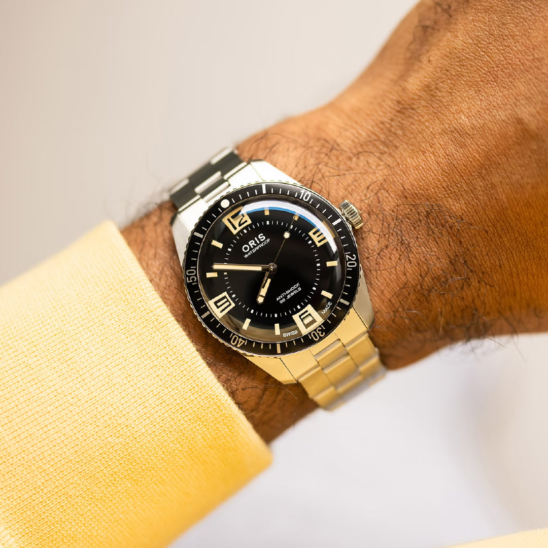 A close-up of a wrist wearing the ORIS Divers 60th Anniversary Edition Calibre 733-1 Automatic 40mm watch, featuring a black dial, bold luminescent numerals, and stainless steel bracelet. The wearer has on a light yellow long-sleeve shirt.