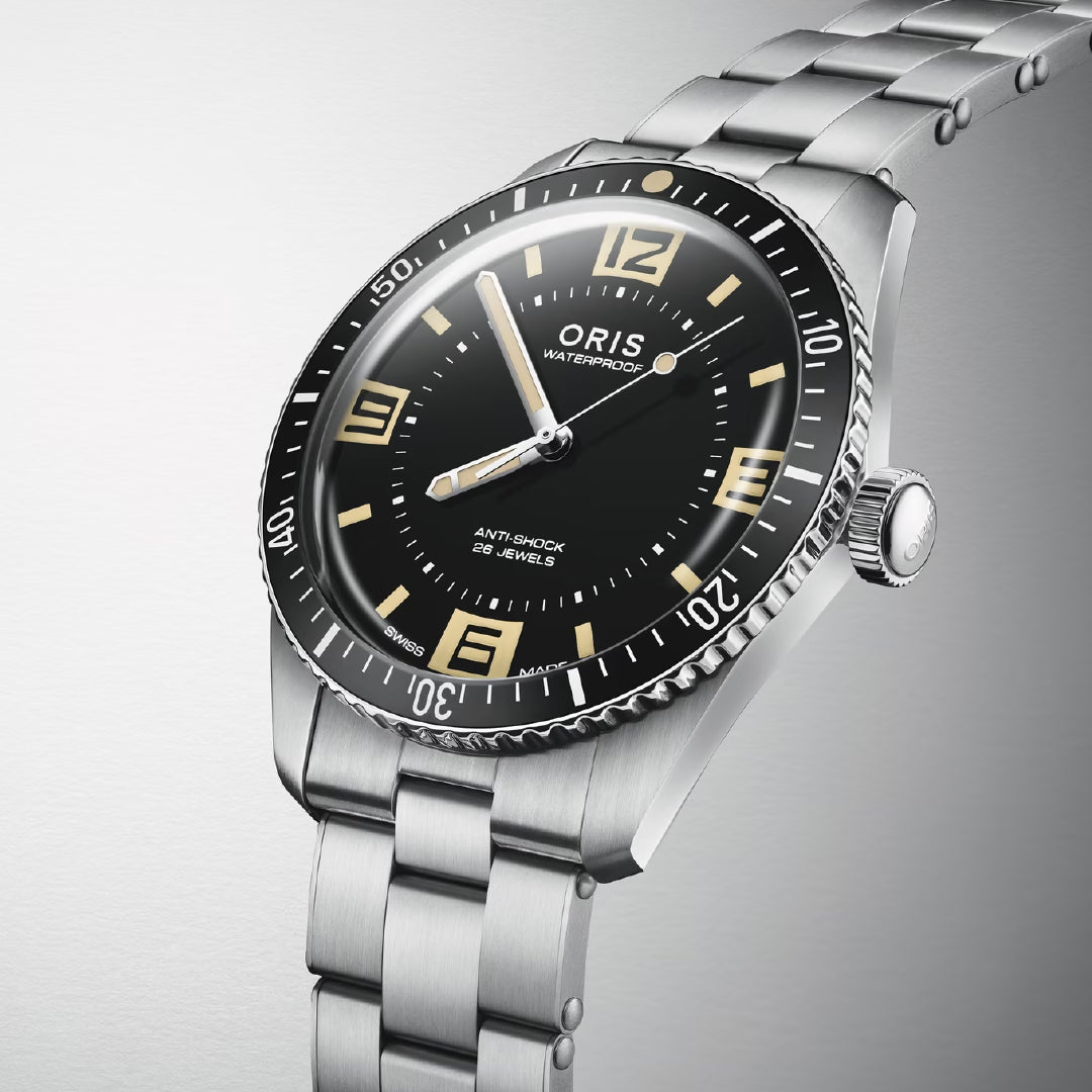 The ORIS Divers 60th Anniversary Edition Calibre 733-1 Automatic Winding 40mm Watch features a stainless steel case, black dial, cream luminescent numerals, and metal bracelet, displayed on a light gray gradient background.