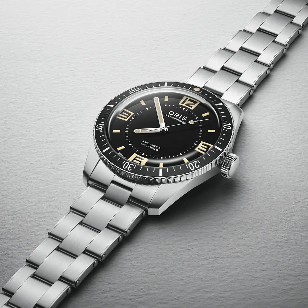 The ORIS Divers 60th Anniversary Edition Calibre 733-1 Automatic 40mm watch features a black dial, silver hour markers, and a stainless steel link band, shown on a light gray textured surface with "ORIS" visible beneath the domed sapphire crystal.