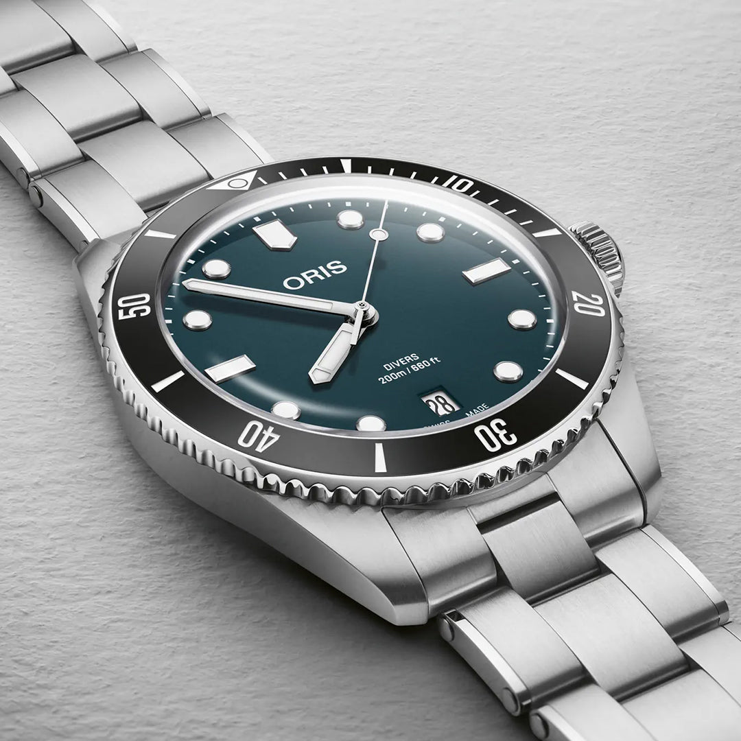 A close-up of the ORIS Divers Date Calibre 733-1 Automatic Winding 39mm watch with a teal dial, luminous markers, and black ceramic bezel rests on a light gray surface. Its brushed stainless steel bracelet showcases Swiss craftsmanship.