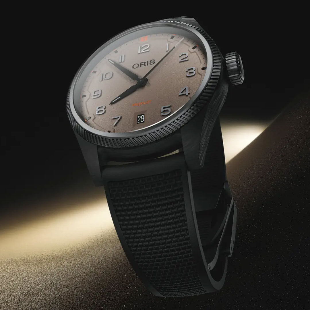 A close-up of the ORIS ProPilot Desert Edition Automatic 41mm shows its brown gradient dial, black numerals, black textured rubber strap, and grey-PVD steel case set against a dark background, with a date window at 6 o’clock.