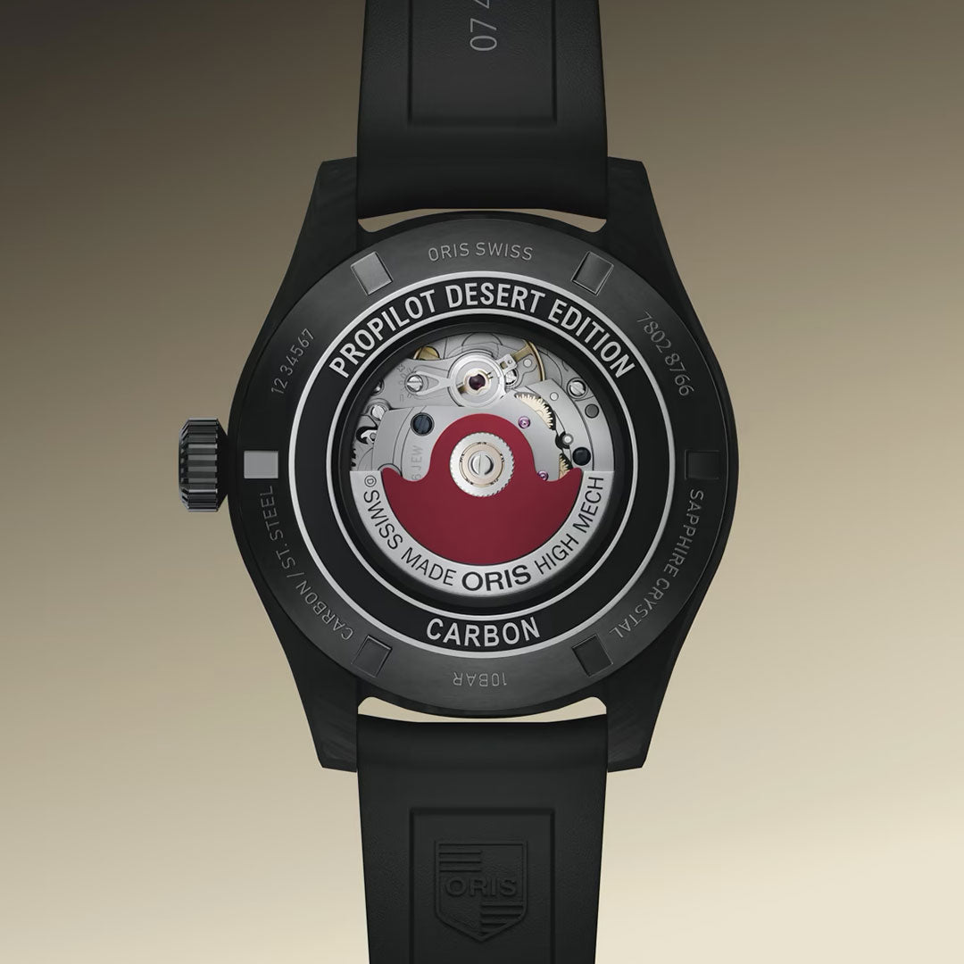 A close-up of the ORIS ProPilot Desert Edition Automatic 41mm Watch shows its exposed mechanical movement and engraved markings on a grey-PVD coated steel case with a black rubber strap.