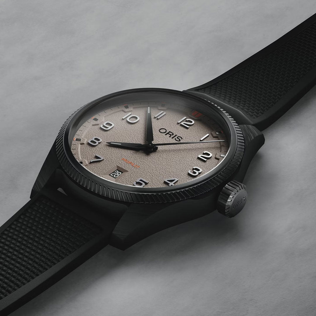 A close-up of the ORIS ProPilot Desert Edition Automatic 41mm watch with a sand beige dial, bold black numerals, black rubber strap, and date at 6 o'clock, displayed on a gray surface.