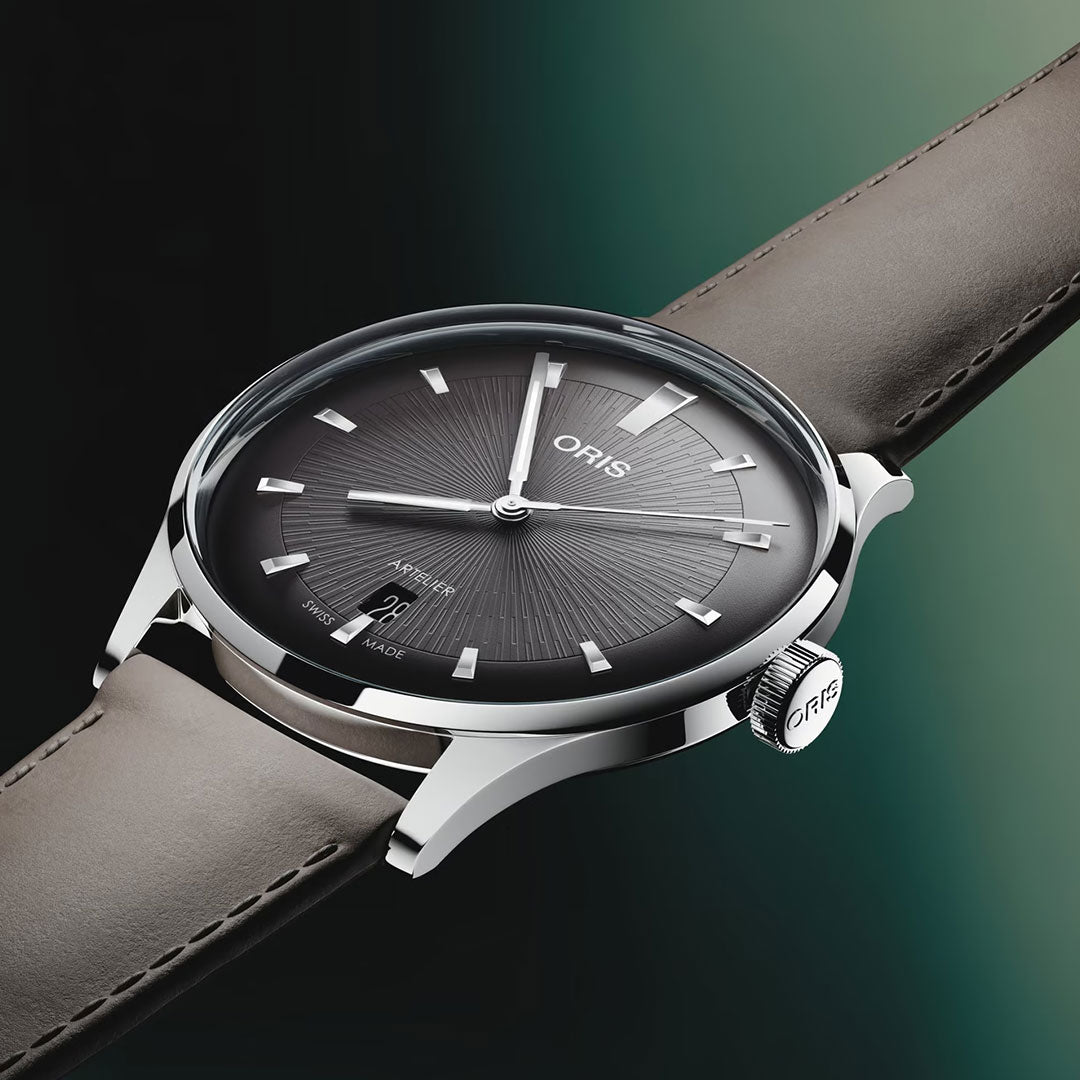 A close-up of the ORIS Artelier Date Calibre 733-1 Automatic 38mm watch, featuring a dark gradient dial, silver markers, date display, and a light brown leather strap by ORIS, set against a black and green gradient background.