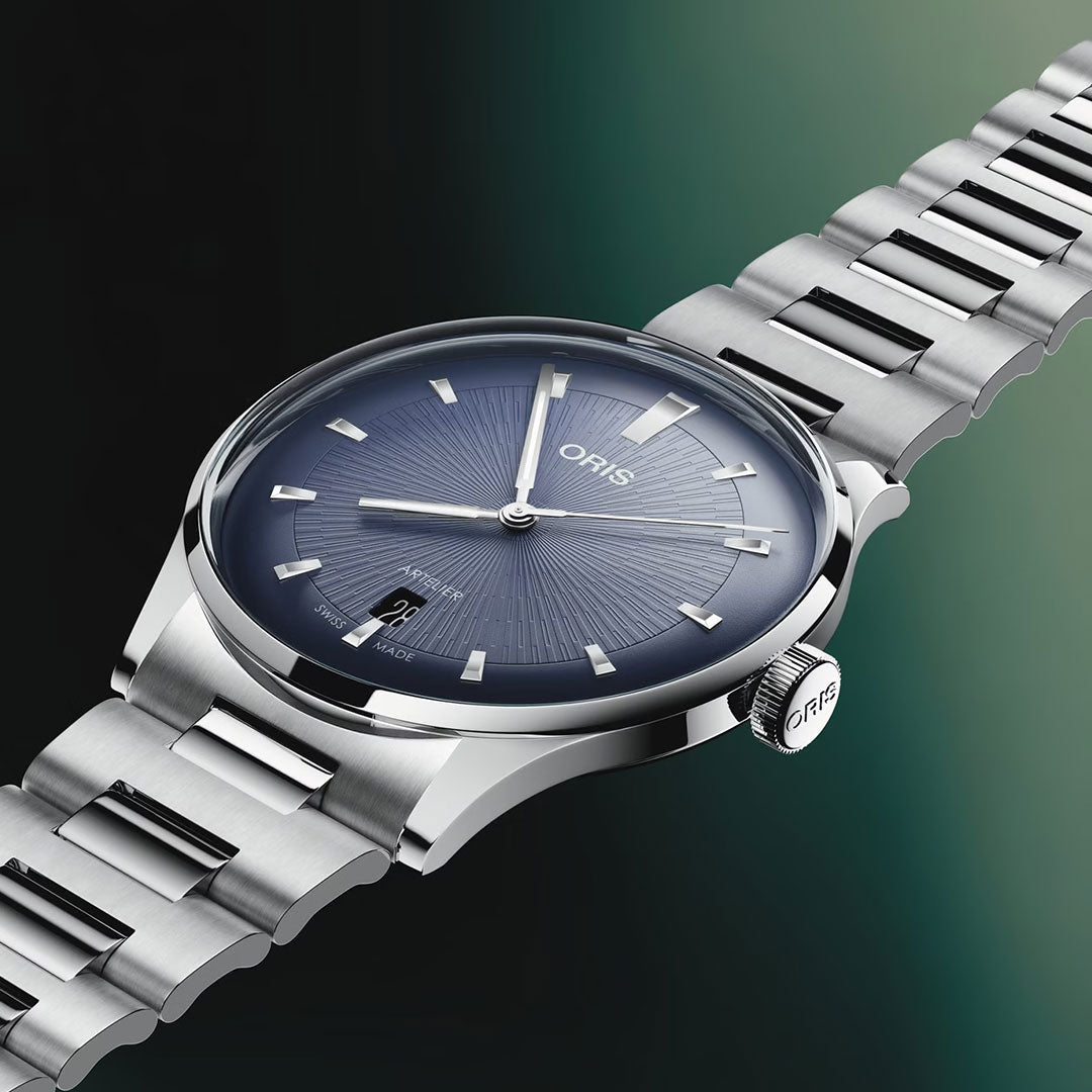 The ORIS Artelier Date Calibre 733-1 Automatic 38mm Watch features a blue sunburst dial with silver hour markers, date at 6 o’clock, and a stainless steel bracelet, displayed on a green-black gradient background.