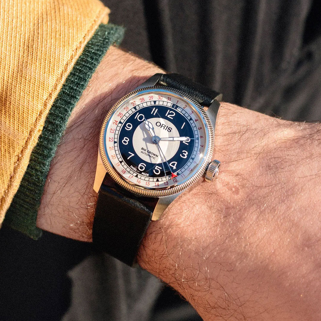 An ORIS Big Crown Pointer Date Bullseye Automatic 38mm watch with a black leather strap, round grey and black dial, red numerals, and silver casing is on the person’s left wrist. The sleeve is yellow with a green cuff.