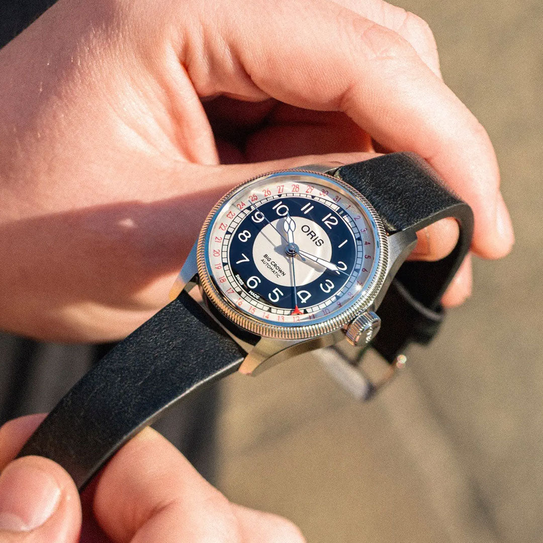 A person holds an ORIS Big Crown Pointer Date Bullseye Automatic 38mm watch with a black leather strap, round dial with white numerals, red and blue bullseye design, silver case, and fluted bezel in natural sunlight.
