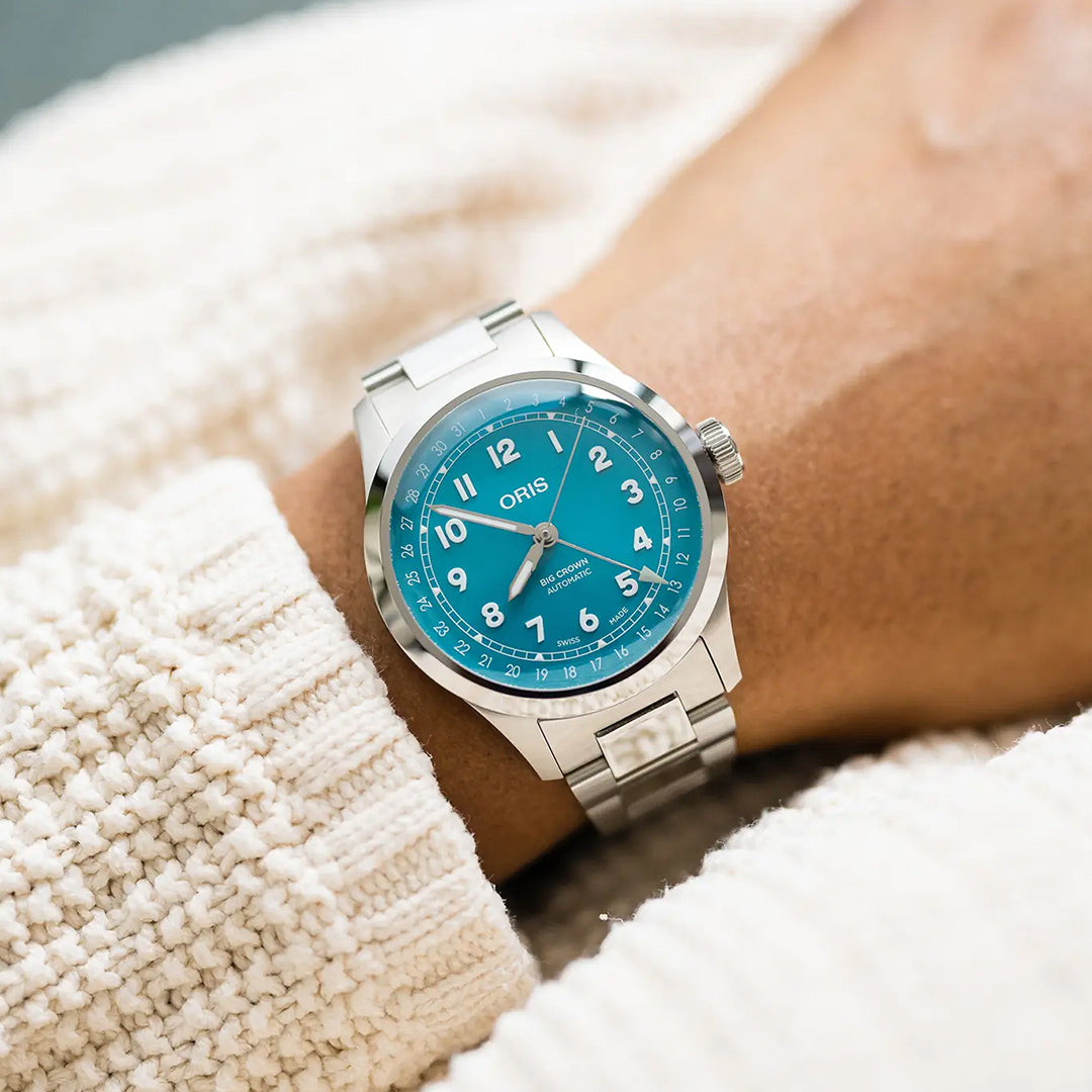 Someone wearing an ORIS Big Crown Pointer Date Calibre 754-1 Automatic 40mm watch with a turquoise blue dial and white numerals, styled with a chunky cream knit sweater.