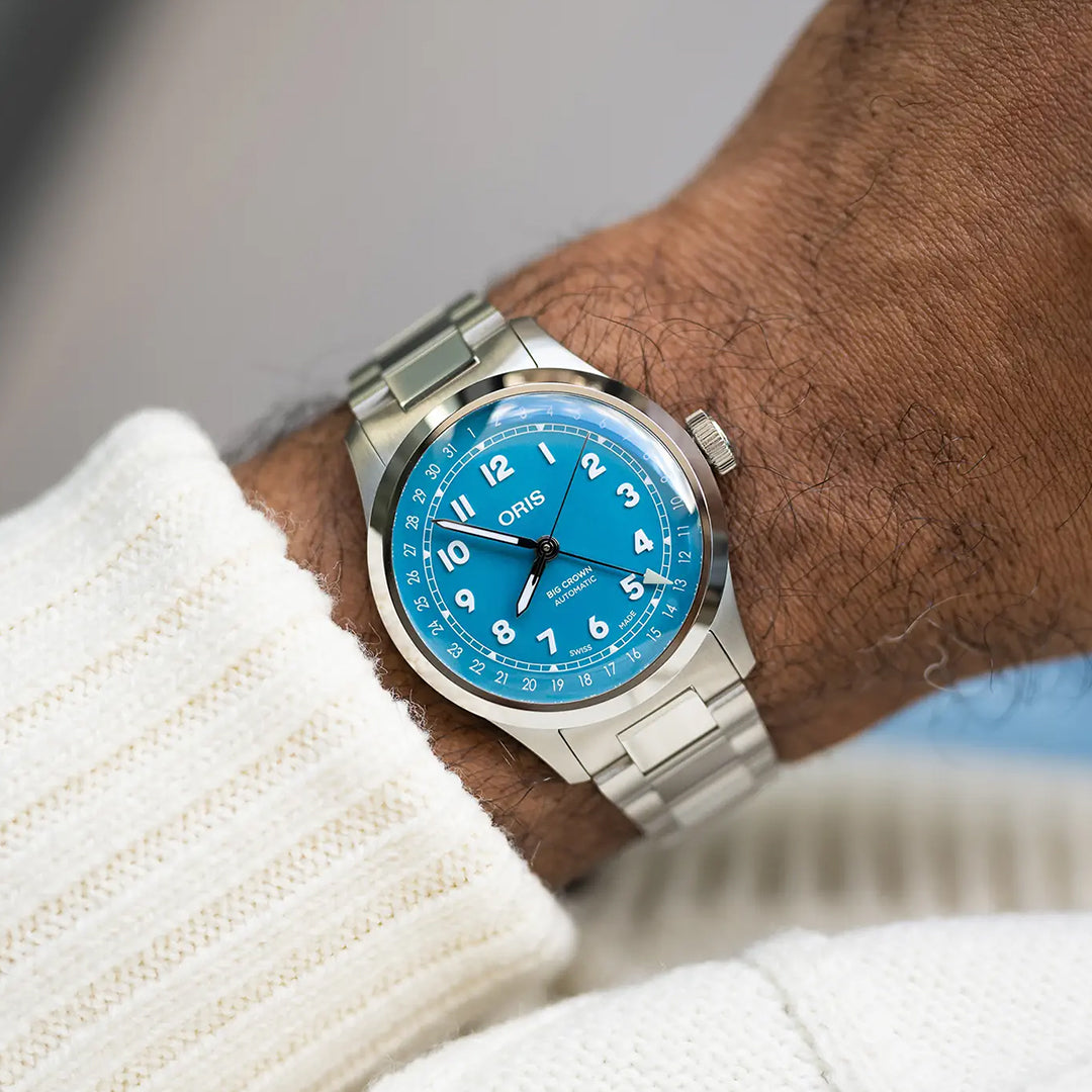 A person wears the ORIS Big Crown Pointer Date Calibre 754-1 Automatic 40mm watch, featuring a stainless steel case, turquoise blue dial with white numerals. They are dressed in a cream-colored ribbed sweater.