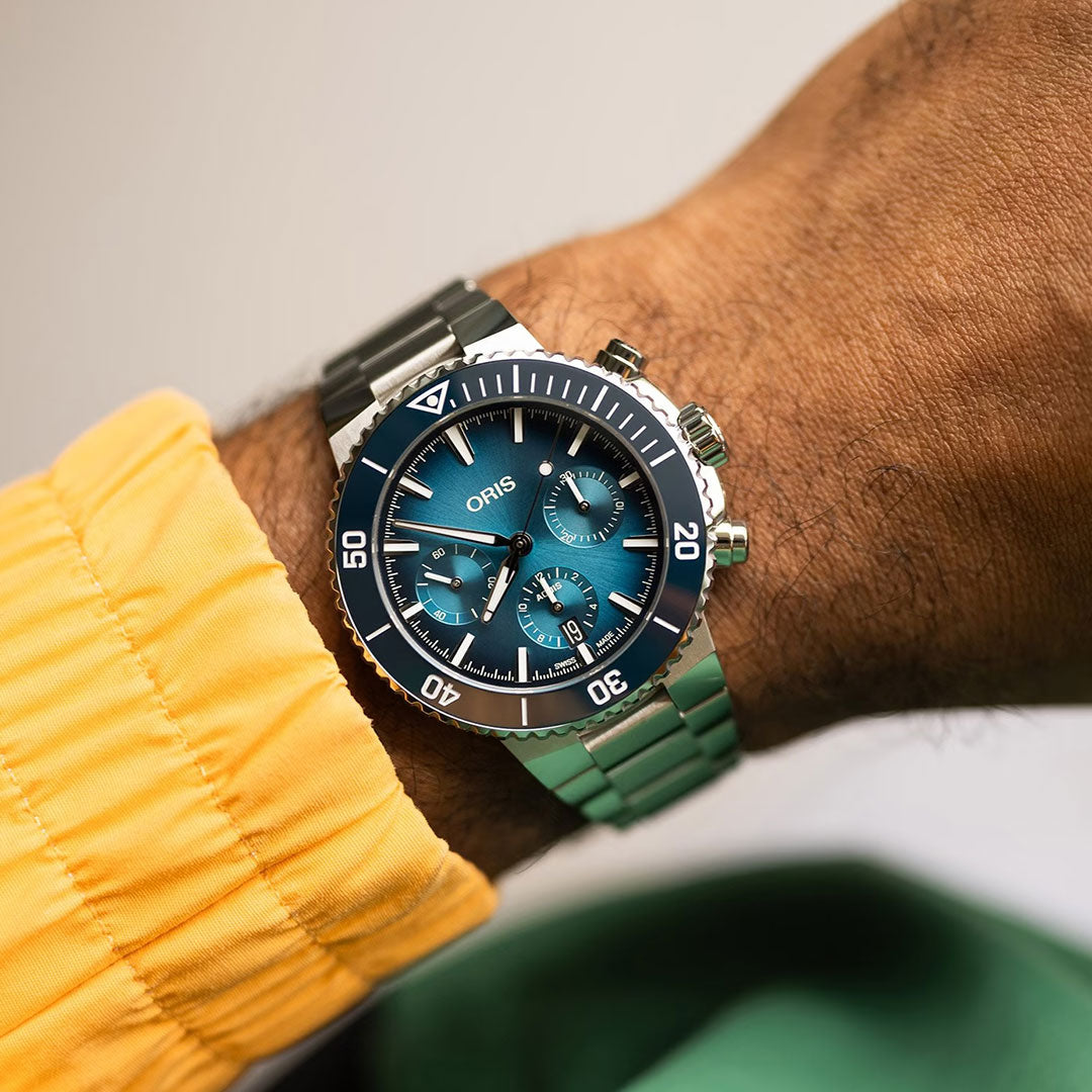 A close-up of a wrist wearing the ORIS Aquis Chronograph Automatic 43.5mm Watch, featuring a silver case, blue dial with three subdials, and a rotating bezel. The toolwatch complements the yellow jacket, showcasing style and performance.