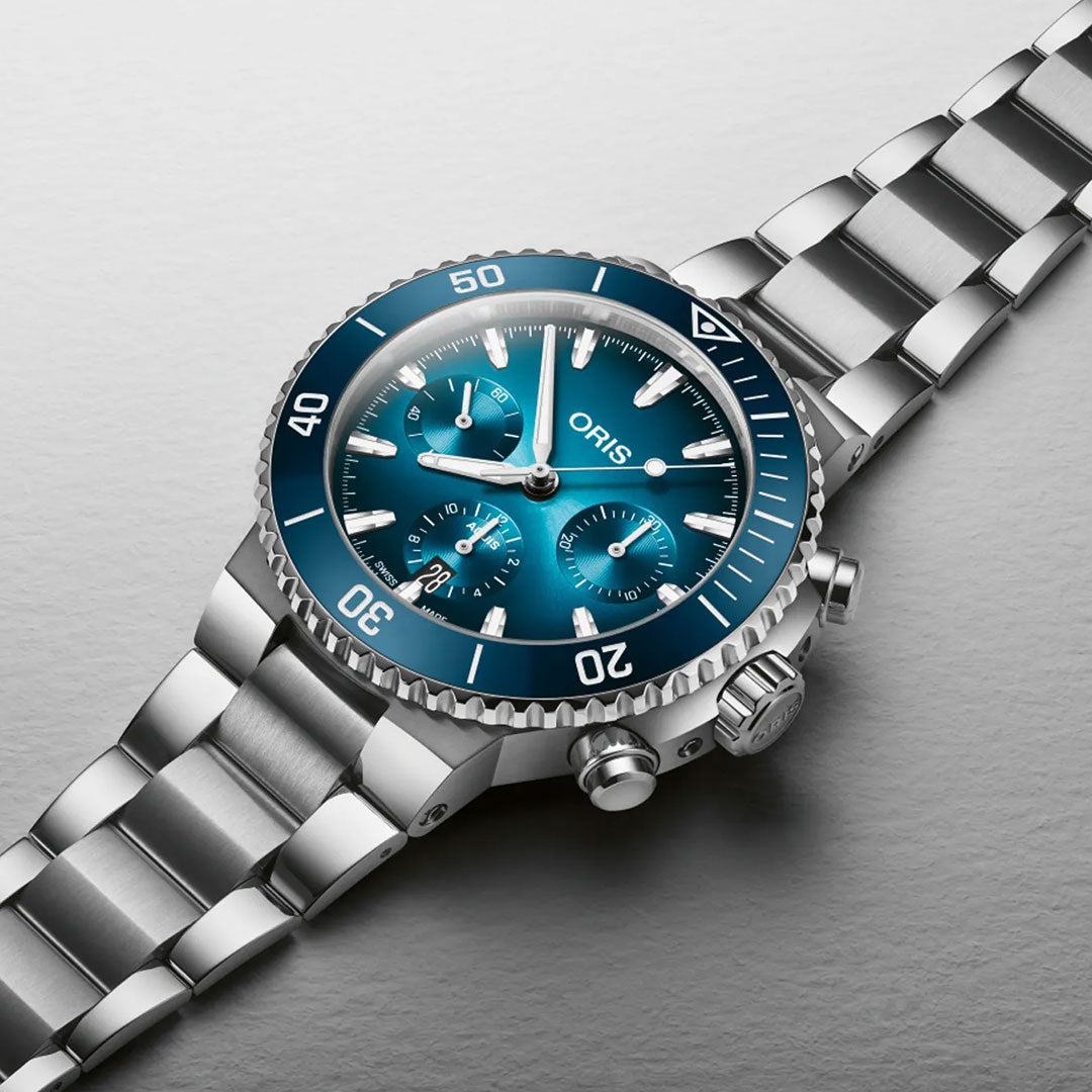 An ORIS Aquis Chronograph Automatic 43.5mm watch with a blue bezel, blue dial, three subdials, and date display lies on a light gray textured surface. The ORIS brand is visible on this high-performance wristwatch.