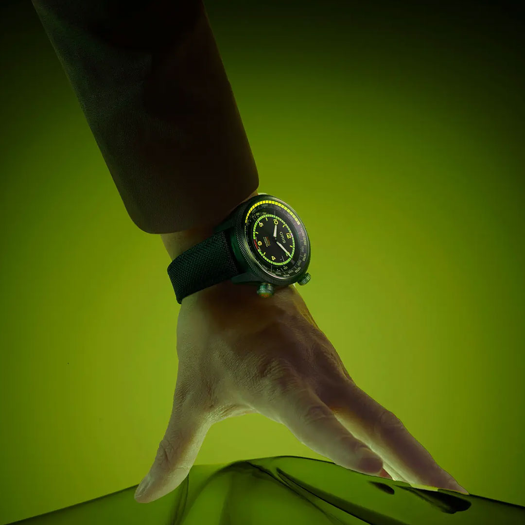 A hand wearing the ORIS Propilot X BWD Limited Edition Automatic Winding 47mm Watch is extended against a glowing green backdrop, fingers softly touching a reflective surface, highlighting its neon green accents.