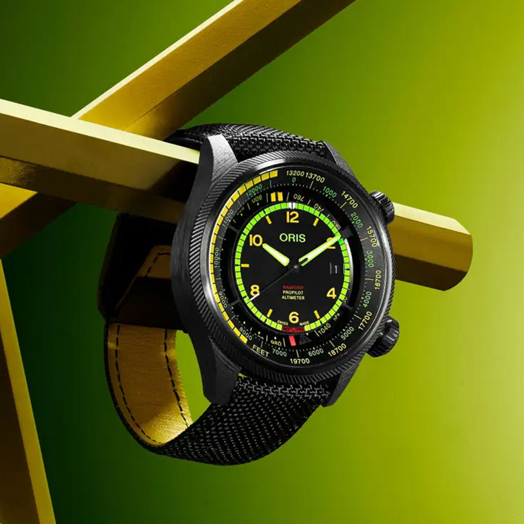 The ORIS Propilot X BWD Limited Edition Automatic 47mm watch features black with yellow and green accents, a black fabric strap, and yellow inner lining, displayed on intersecting yellow beams against a vibrant green background.
