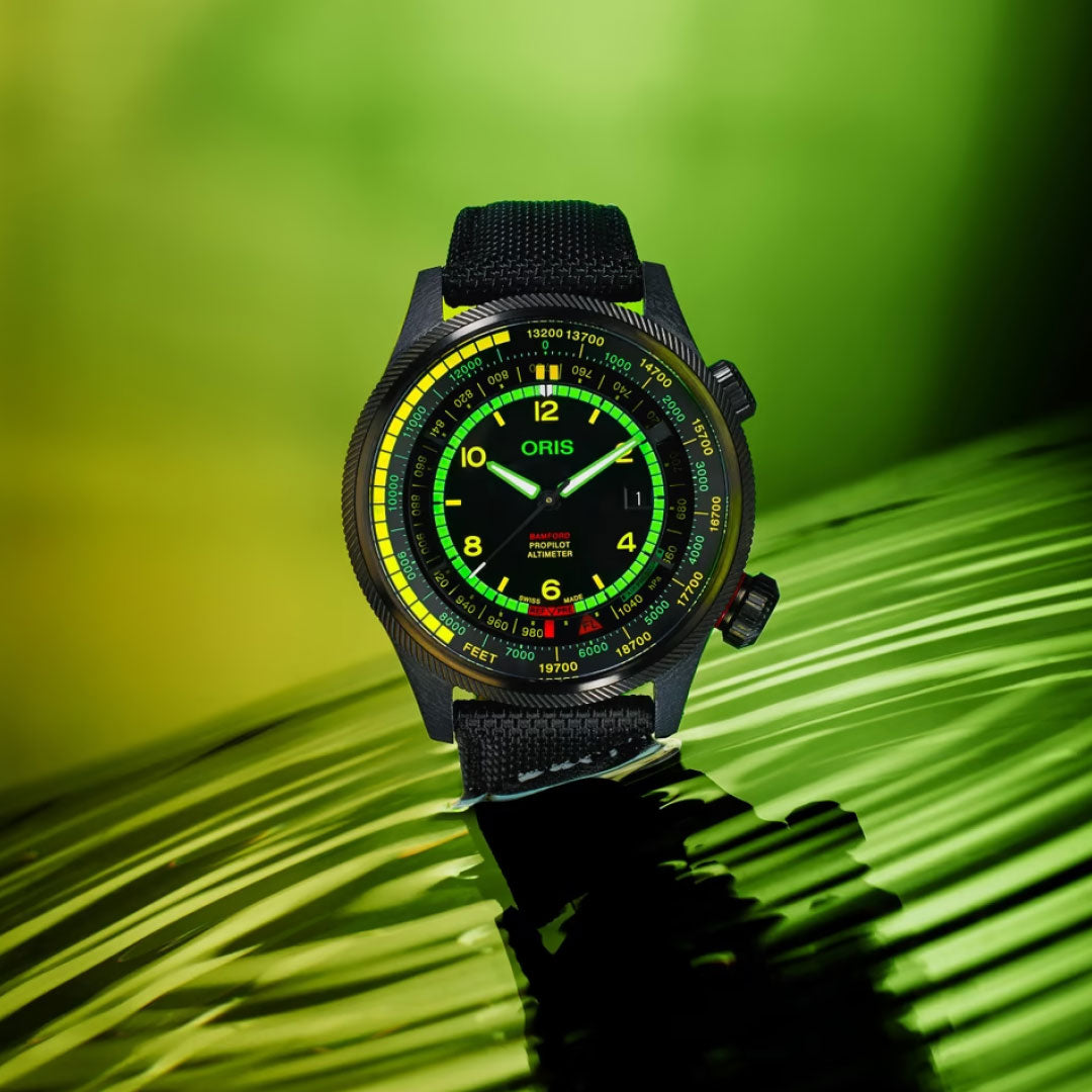 The ORIS Propilot X BWD Limited Edition Automatic Winding 47mm Watch is shown on a palm leaf, with a glowing green and yellow dial, black fabric strap, dual crowns, and striking Limited Edition appeal against a bright green gradient backdrop.