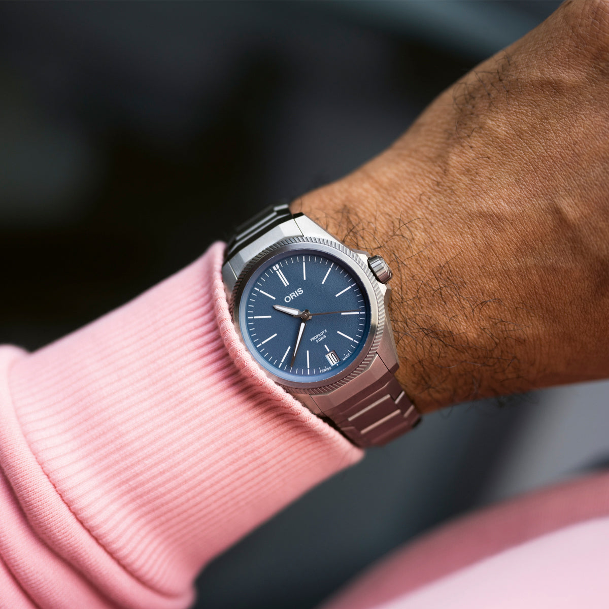 A person wearing an ORIS Propilot X Calibre 400 Titanium Automatic Winding 39mm watch with a blue dial and silver band. The watch, which highlights the Calibre 400 movement, displays the brand name "ORIS." The person is dressed in a pink ribbed long-sleeve shirt.