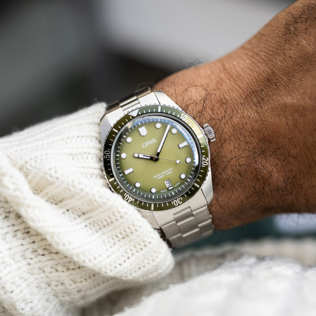 A person's wrist adorned with an ORIS Divers Sixty-Five Date Automatic 40mm watch, showcasing a green dial, unidirectional bezel, and luminescent markers, is partially covered by a cream knitted sweater.