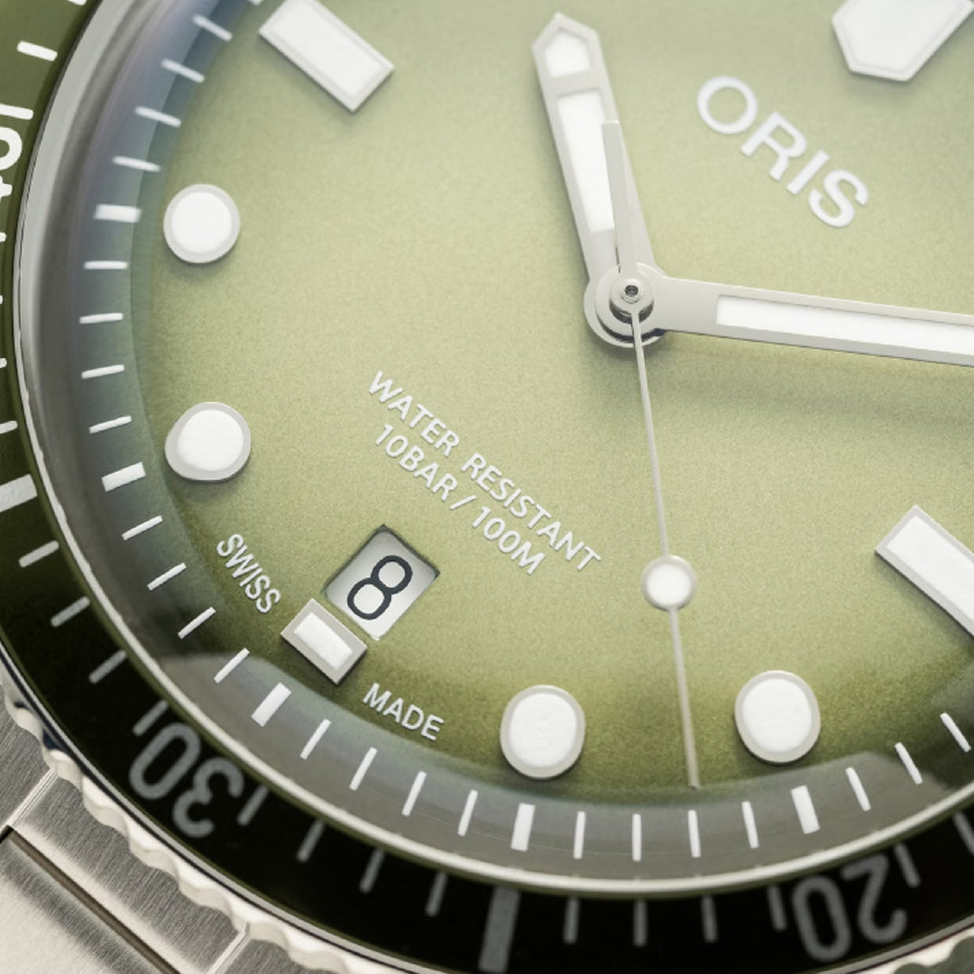 A close-up reveals the green dial of the Oris Divers Sixty-Five Date Automatic Winding 40mm Watch. It features luminous white hour markers, hands, and a date window showing "8" in a stainless steel case. The bezel is marked up to 30 with "Oris," "Water Resistant 10BAR/100M," and "Swiss Made.