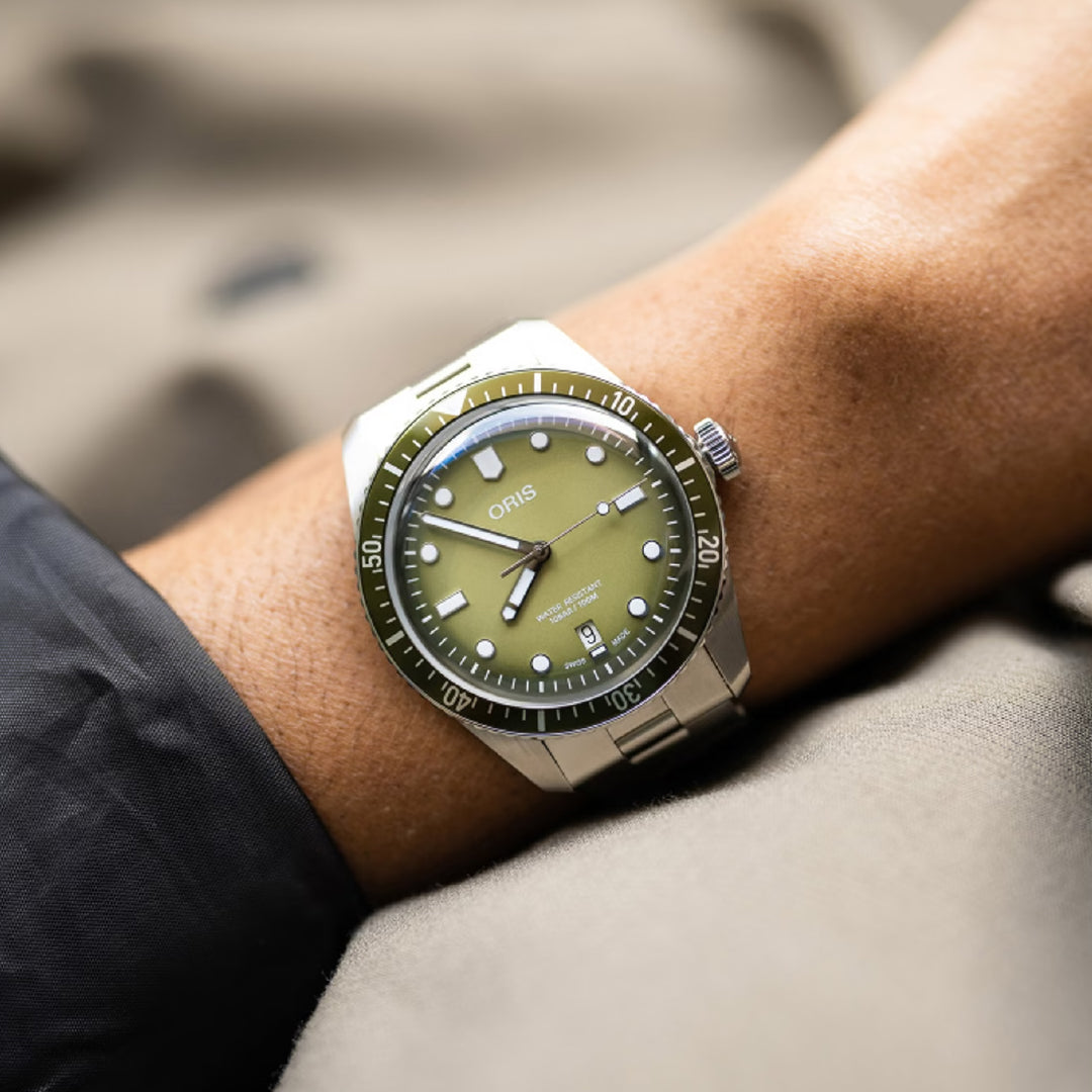 A person wears an Oris Divers Sixty-Five Date Automatic Winding 40mm watch with a striking green dial, white markers, black rotating bezel, and silver bracelet. The ORIS watch gracefully sits over a dark sleeve against a blurred background.