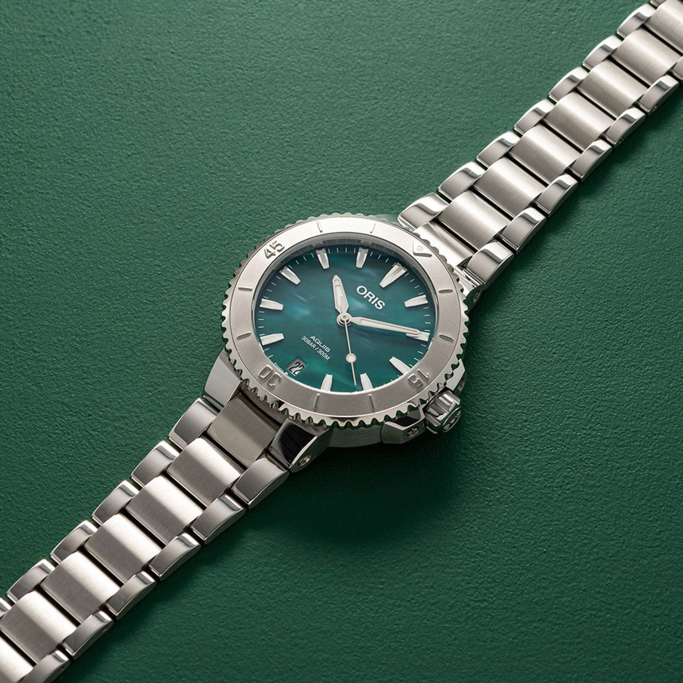 The Oris Aquis X Bracenet Automatic 36.5mm Watch by ORIS is a sophisticated stainless steel timepiece that showcases a teal dial and date display on its metal link bracelet, set against a green backdrop. Silver hands and markers enhance the visible branding on the dial, making it an elegant accessory for any occasion.