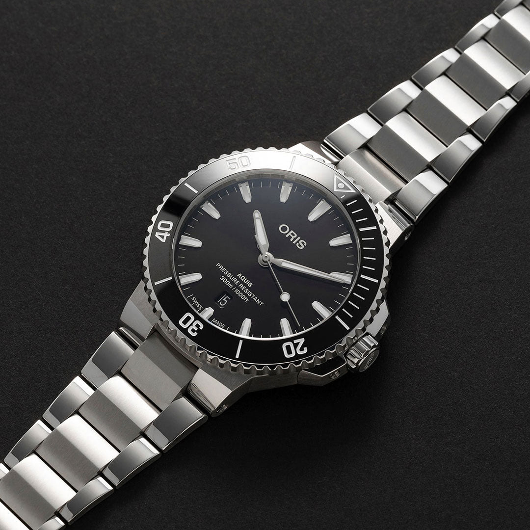 This ORIS Aquis Date Automatic Winding 43.5mm Watch is a stylish silver stainless steel timepiece with a black dial, featuring white hands and hour markers. It includes a rotating bezel with minute markings, impressive water resistance, and a date display located at 6 o'clock, all set against a dark backdrop.