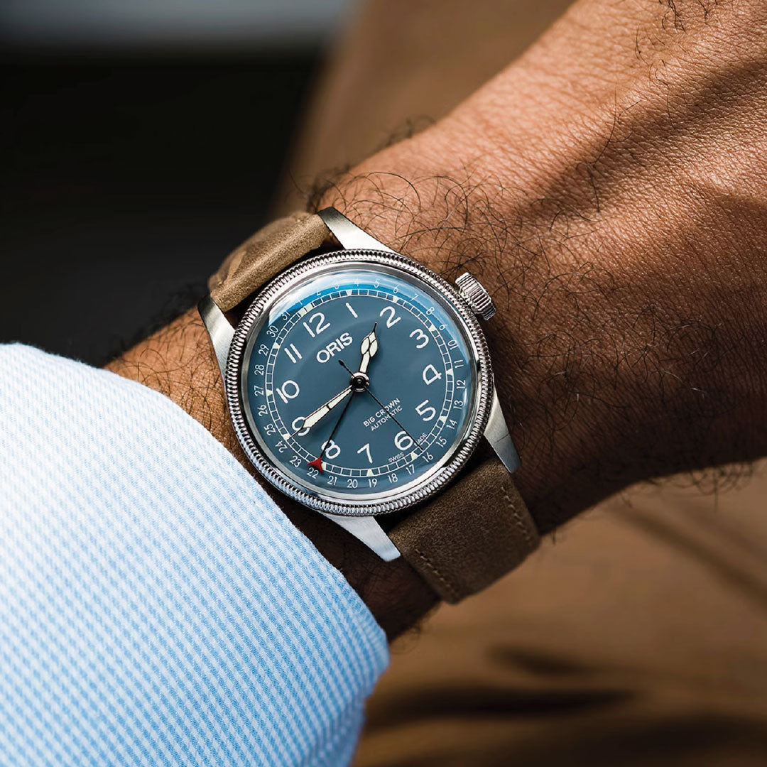 A close-up of a wrist wearing the ORIS Big Crown Pointer Date Calibre 754 Automatic 40mm watch with a blue dial, white numerals and hands, and a brown leather strap. The person is dressed in a light blue shirt.